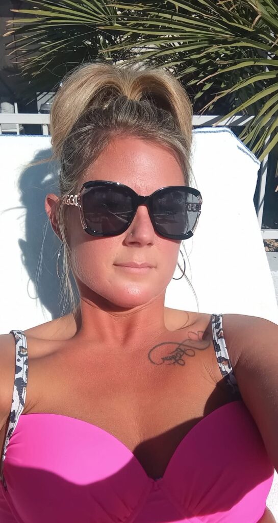 Nikki relaxing poolside wearing sunglasses and a pink swimsuit.